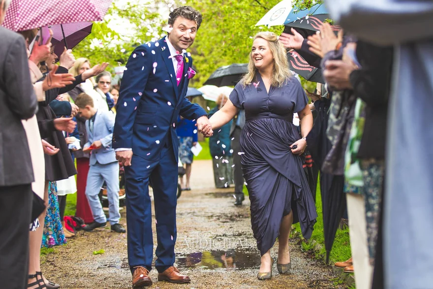 A couple walking hand in hand through confetti and guests with umbrellas